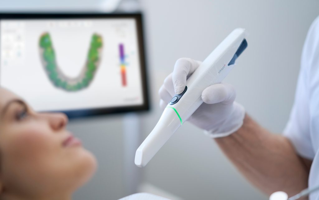 Dentist scanning patient's teeth with 3Shape Trios 5 intraoral scanner, with digital scan visible on computer.